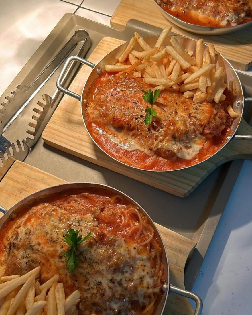 Duas frigideiras de parmegiana na bancada, uma com fritas e outra com espaguete